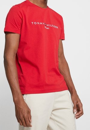 Mand iført en rød Tommy Hilfiger T-shirt med logotekst og off-white bukser, stående mod en ensartet lys baggrund.