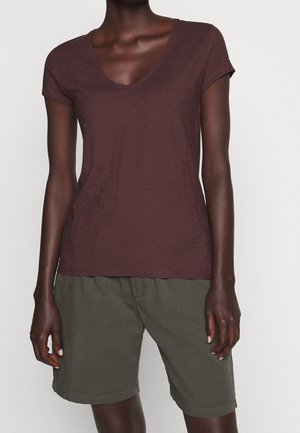 Personne à la peau foncée portant un T-shirt marron à manches courtes et col en V et un short vert olive arrivant aux genoux, debout devant un fond uni.