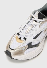 White athletic shoe with mesh upper, gold and silver accents, black detailing, and round toe. Features a textured sole and laces for closure.