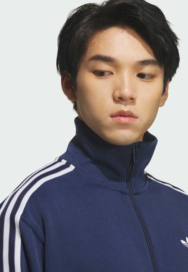 Giovane uomo con capelli neri corti che indossa una giacca blu navy con zip e strisce bianche sulle spalle, che guarda in basso verso sinistra.