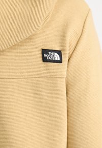 Beżowy polarowy hoodie z teksturowanym, prążkowanym wzorem oraz czarną naszywką z logo na lewej piersi.