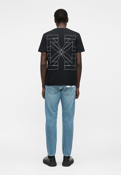 Άτομο που στέκεται με την πλάτη προς τα εμπρός, φοράει μαύρο μπλουζάκι με λευκά γεωμετρικά βέλη, φωτοστέφανο ανοιχτό μπλε τζιν και μαύρα παπούτσια σε λευκό φόντο.