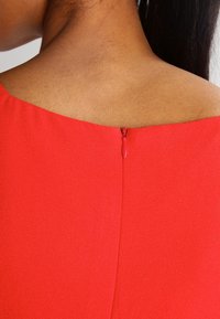 Červené šaty s hladkou tkanou texturou, skrytým zipem na zadní straně a širokým výstřihem, které zdůrazňují čistý a upravený design.