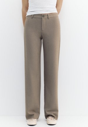 Taupe nohavice rovného strihu nosené s bielym topom a béžovými šnurovacími topánkami, stojace pred jednoduchým bielym pozadím.