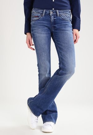 Osoba w niebieskich jeansach o prostych nogawkach i białych trampkach, stojąca ze skrzyżowanymi nogami, częściowo widoczna granatowa koszula.