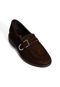 Mocassin en daim marron foncé avec boucle argentée et semelle noire texturée, vue en angle sur fond blanc.