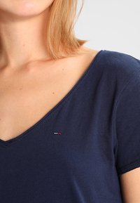 Žena v námořnicky modrém T-tričku s v�ýstřihem do V, na hrudi poblíž klíční kosti má malé obdélníkové logo v červené, bílé a modré barvě.