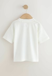 T-shirt blanc à manches courtes et col rond suspendu sur un cintre en bois contre un mur clair uni.