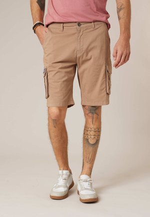 Homme portant un short cargo beige, des baskets blanches et une chemise rose, montrant des jambes tatouées et des bracelets au poignet gauche.