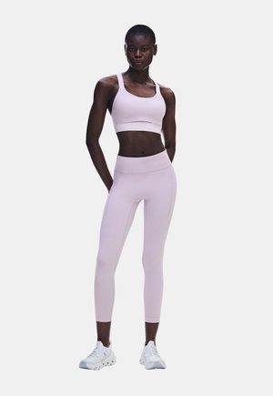 Frau steht in einem helllila Sport-BH und passenden Leggings mit weißen Sneakers vor einem schlichten, hellen Hintergrund, die Hände hinter dem Rücken.