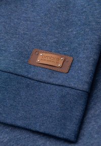 Tejido azul con una textura jaspeada, que presenta una etiqueta de cuero marrón con remaches plateados, nombre de la marca grabado, detalle texturizado en los bordes.