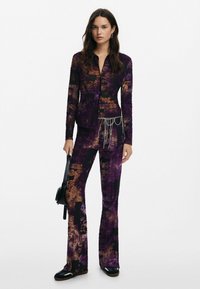 Outfit a motivi viola e neri con top aderente a maniche lunghe e pantaloni a zampa, caratterizzato da un accento a catena in vita. Tessuto morbido.