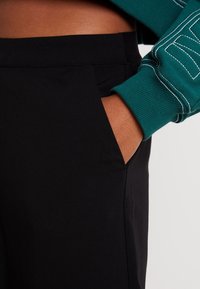 Svarta tygbyxor med en slät textur, med sidfickor och kontrastsömnadsdetaljer, tillsammans med en grön sweatshirtärm.