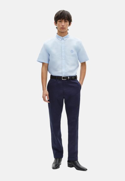 Camisa corta de manga azul claro con cierre de botones, logo en el pecho; combinada con pantalones oscuros de color navy y zapatos de cuero negro, con una textura suave.