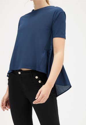 Femme portant un haut asymétrique bleu marine à manches courtes avec un dos évasé et un pantalon noir taille haute à boutons décoratifs.