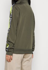 Zielona kurtka z suwakiem, z czarnymi i neonowymi akcentami na rękawach oraz wysokim kołnierzem. Wykonana z gładkiego, sportowego materiału z ściągaczami przy rękawach.