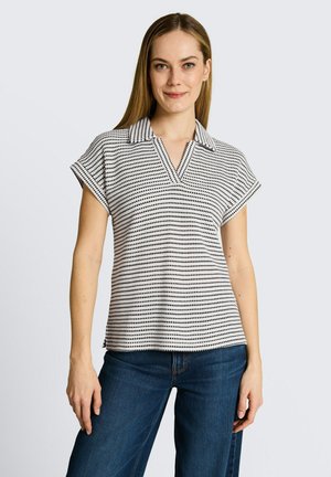 Kobieta w koszuli z krótkim rękawem i kołnierzykiem w czarno-białe poziome kropkowane paski oraz w niebieskich dżinsach, stojąca na jednolitym tle.