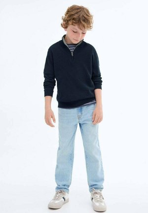 Navy fleece zip-up trui met een lichtgrijze kraag, gecombineerd met lichtblauwe jeans en grijze sneakers met donkere accenten.