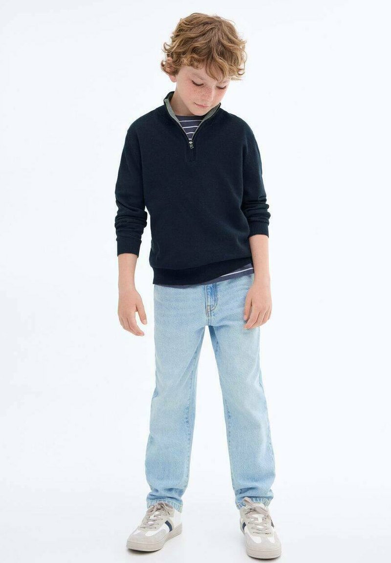 Marinblå fleece zip-up-tröja med en ljusgrå krage, kombinerad med ljusblå jeans och grå sneakers med mörka accenter.