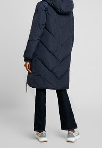 Marineblauwe gewatteerde jas met een capuchon, voorzien van een chevron-quilt patroon. Draagt over een donkere spijkerbroek en gecombineerd met multicolor sneakers.