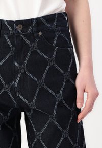 Mørke denimjeans med et geometrisk mønster i lys blå tråd. Stoffet har en tekstur, med høy midje-design og metallbeslag.
