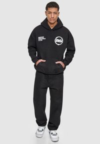 Svart hettegenser med lommer foran og grafisk trykk, kombinert med svarte bukser og hvite sneakers med svarte detaljer. Avslappet passform.