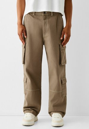 Proste khaki spodnie typu cargo z klapkowymi kieszeniami na udach i łydkach, noszone z białymi sneakersami i białym przyciętym topem.