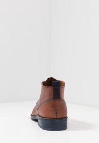 Bottine en cuir marron avec une texture lisse, accents latéraux élastiques bleu marine et une semelle noire contrastante. Détails de couture visibles à l'arrière.