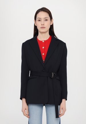 Navy tailored blazer with a belt, featuring a notch lapel and two front pockets, layered over a red ribbed top with gold buttons.