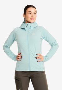 Kvinna i ljusblå zip-up huvtröja och mörkbruna byxor, stående med händerna på höfterna, tittar åt höger.