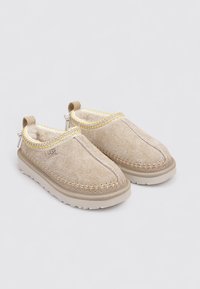 Une paire de pantoufles UGG beiges à enfiler avec doublure en laine polaire, détails cousus et semelles en caoutchouc structuré sur un fond clair.