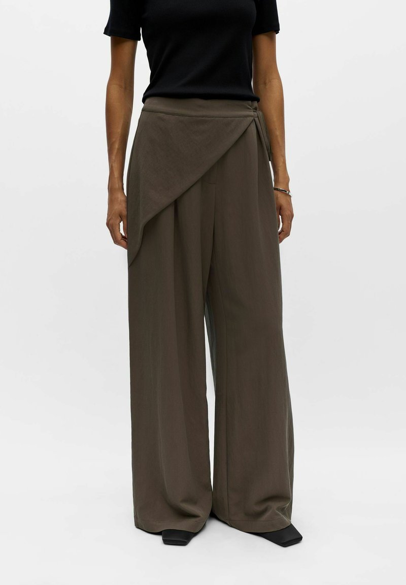 Femme portant un pantalon large vert olive avec un détail asymétrique en forme de drapé sur un haut noir à manches courtes et des chaussures noires.