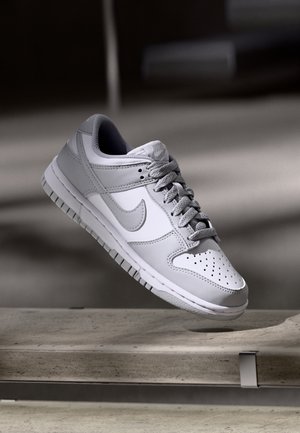Witte en grijze Nike-sneaker met veters, zwevend boven een betonnen oppervlak tegen een wazige donkere achtergrond.
