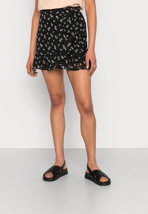 Schwarzer, floraler Wickelrock mit rüschengesäumtem Saum, aus leichtem Stoff gefertigt. Kombiniert mit schwarzen Sandalen, die ein überkreuztes Design der Riemen aufweisen.
