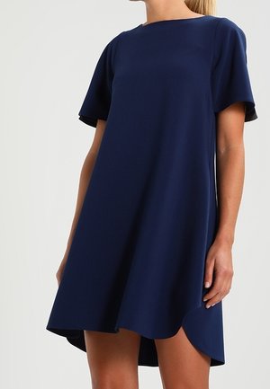 Mujer que lleva un vestido azul marino de manga corta, corte en línea A, con escote redondeado y un dobladillo ligeramente más largo en la parte trasera.
