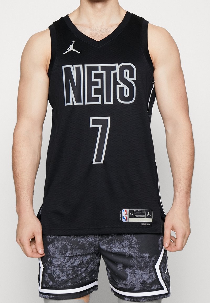 Homme portant un maillot de basket noir des Brooklyn Nets numéro 7 et un short noir assorti à motifs, debout sur un fond uni.