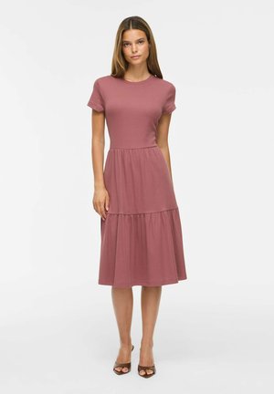 Jeune femme portant une robe midi manches courtes couleur vieux rose avec une jupe à volants et des talons ouverts marron, debout devant un fond blanc.