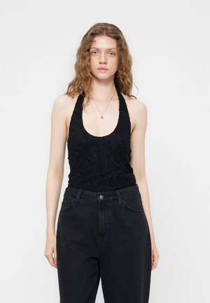 Νεαρή γυναίκα με κυματιστά μαλλιά που φοράει μαύρο υφασμάτινο τοπ χωρίς μανίκια με δέσιμο στον λαιμό και μαύρο τζιν ψηλόμεσο μπροστά σε μονόχρωμο φόντο.