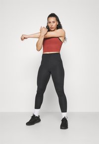Rød kort tanktop og høytlivede sorte leggings. Modellen strekker armene, iført svarte joggesko med hvite detaljer og sokker. Enfarget bakgrunn.