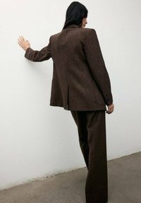 Blazer in lana marrone con una vestibilità rilassata, caratterizzato da uno spacco sul retro, indossato con pantaloni abbinati. Texture liscia e design su misura.