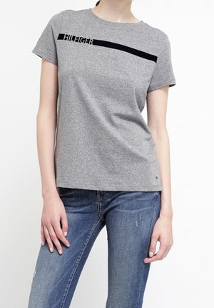 Vrouw draagt een grijs T-shirt met korte mouwen, voorzien van een zwarte diagonale streep en logo, gecombineerd met blauwe jeans met slijtageplekken.