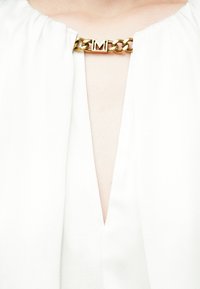 Haut blanc à encolure plongeante en V, avec un accent de chaîne dorée ornée d'une lettre "M" embossée au col. Tissu doux, légèrement texturé.