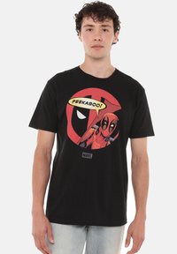 Young man wearing a black Marvel Deadpool t-shirt with Deadpool character and speech bubble saying "Peekaboo!" on front, paired with light jeans.