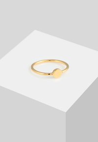 Inel aurit cu un band subțire și un vârf rotund, neted. Design minimalist cu o finisare lucioasă, așezat pe o suprafață geometrică gri.