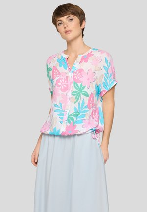 Frau trägt eine kurzärmelige Bluse mit Blumenmuster in Pink, Blau, Grün und Beige, kombiniert mit einem hellblauen Rock vor einem einfarbigen Hintergrund.