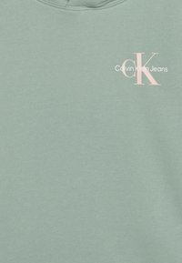 Sudadera verde claro de textura suave, con un logo impreso en rosa y blanco. Cuello redondo con ribetes.