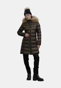 Cappotto imbottito marrone con cappuccio in pelliccia sintetica, dotato di chiusura con zip e tasche laterali, indossato con pantaloni neri e stivali neri con tacco.