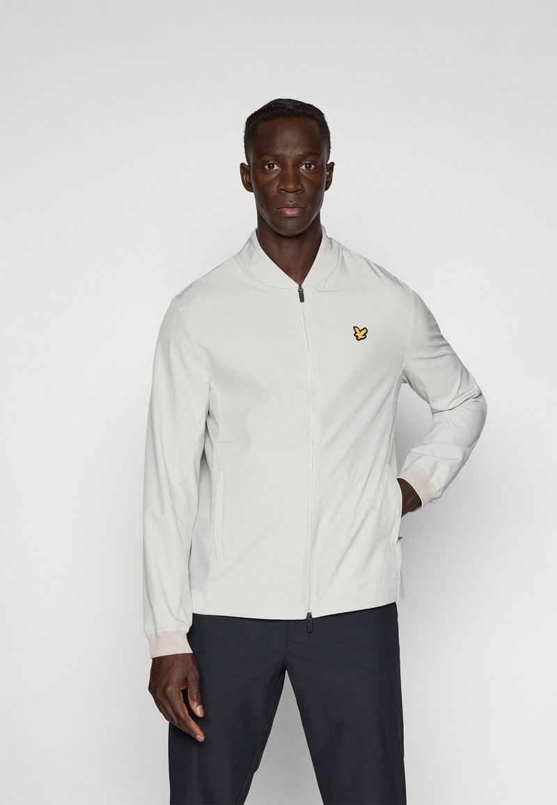Veste bomber gris clair avec fermeture éclair à l'avant, poignets élastiques et poches latérales. Présente un petit logo jaune sur la poitrine.