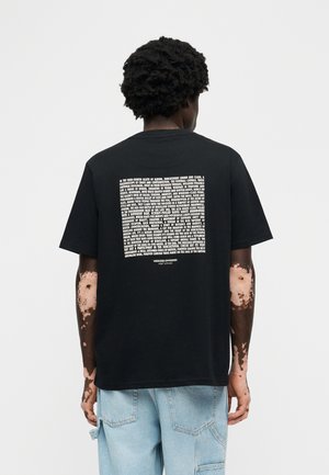 Άνδρας με λεύκη στα χέρια, φορόντας μαύρο μπλουζάκι με λευκό κείμενο και ανοιχτό μπλε τζιν, στέκεται με την πλάτη προς το μέρος μας μπροστά σε απλό φόντο.