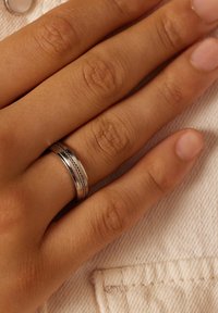 Zilveren bandring met een centrale getextureerde sectie en gladde buitenranden, gedragen op een hand met licht getextureerde huid tegen een neutrale achtergrond.